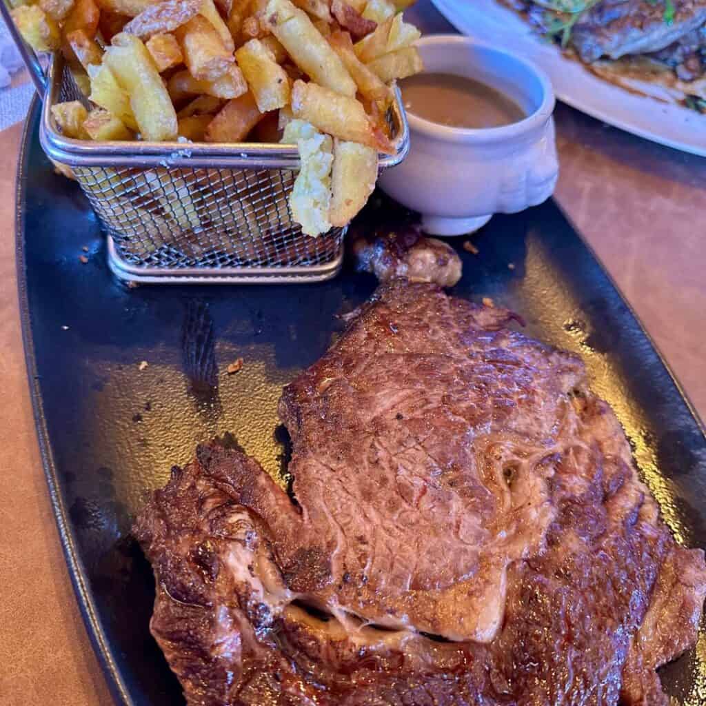 Juicy grilled steak served with a small metal basket of crispy golden fries and a pot of creamy sauce on a black plate.