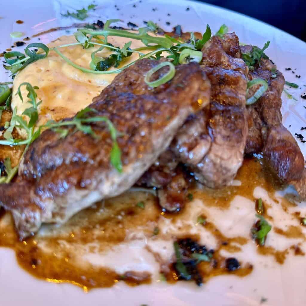 Sliced seared meat served in a rich sauce with creamy mashed potatoes, topped with fresh herbs and spring onion garnish.