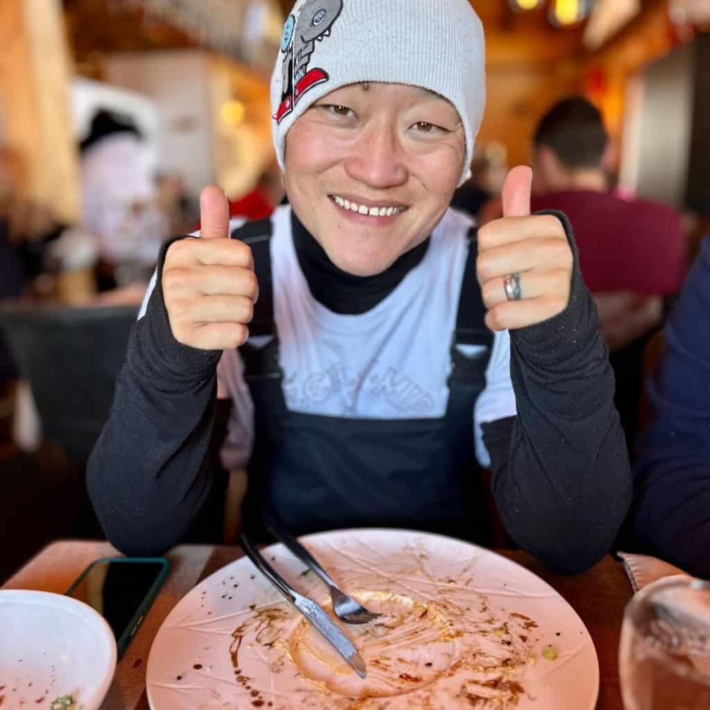 A smiling person wearing a beanie with a cartoon design gives two thumbs up while sitting at a restaurant table with an empty plate, clearly satisfied after a meal.