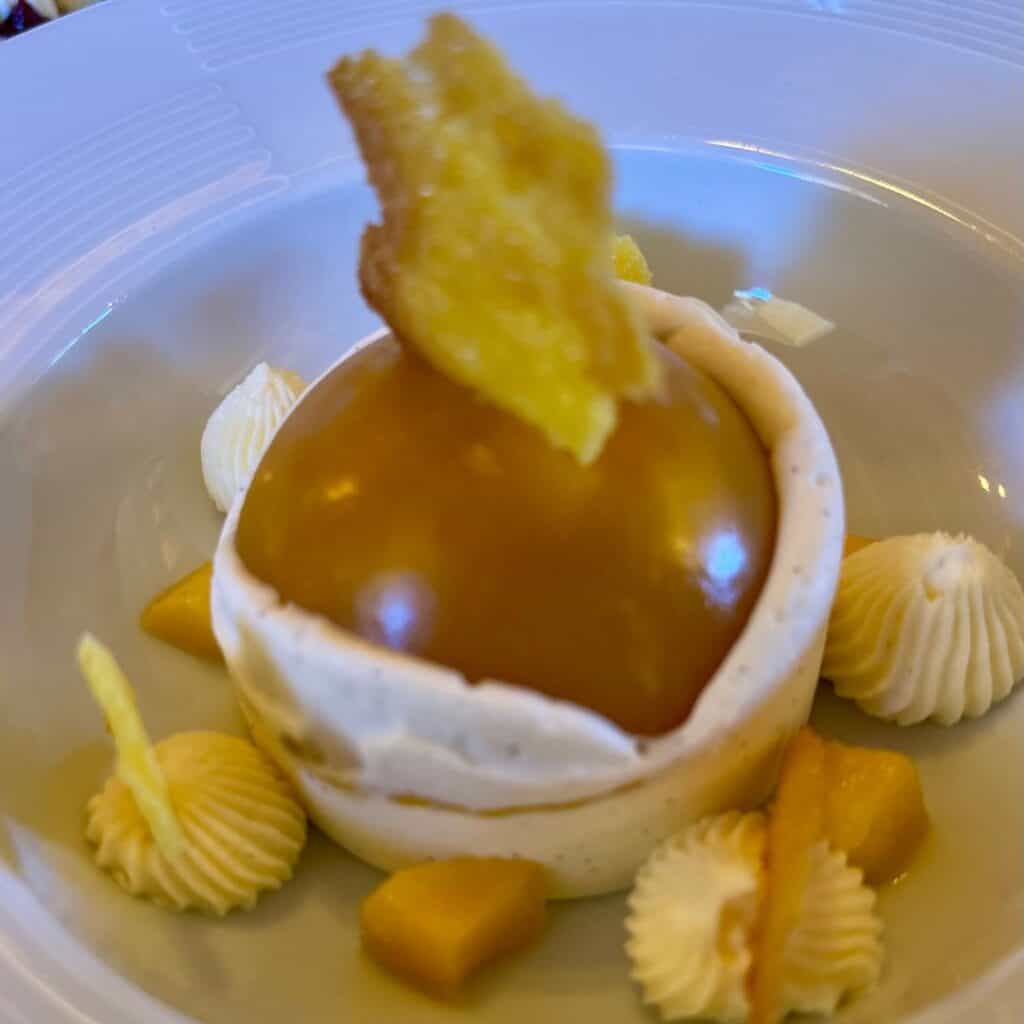 Modern plated dessert with a glossy golden dome surrounded by piped cream, diced fruit, and a crisp garnish, sitting in a shallow white bowl.