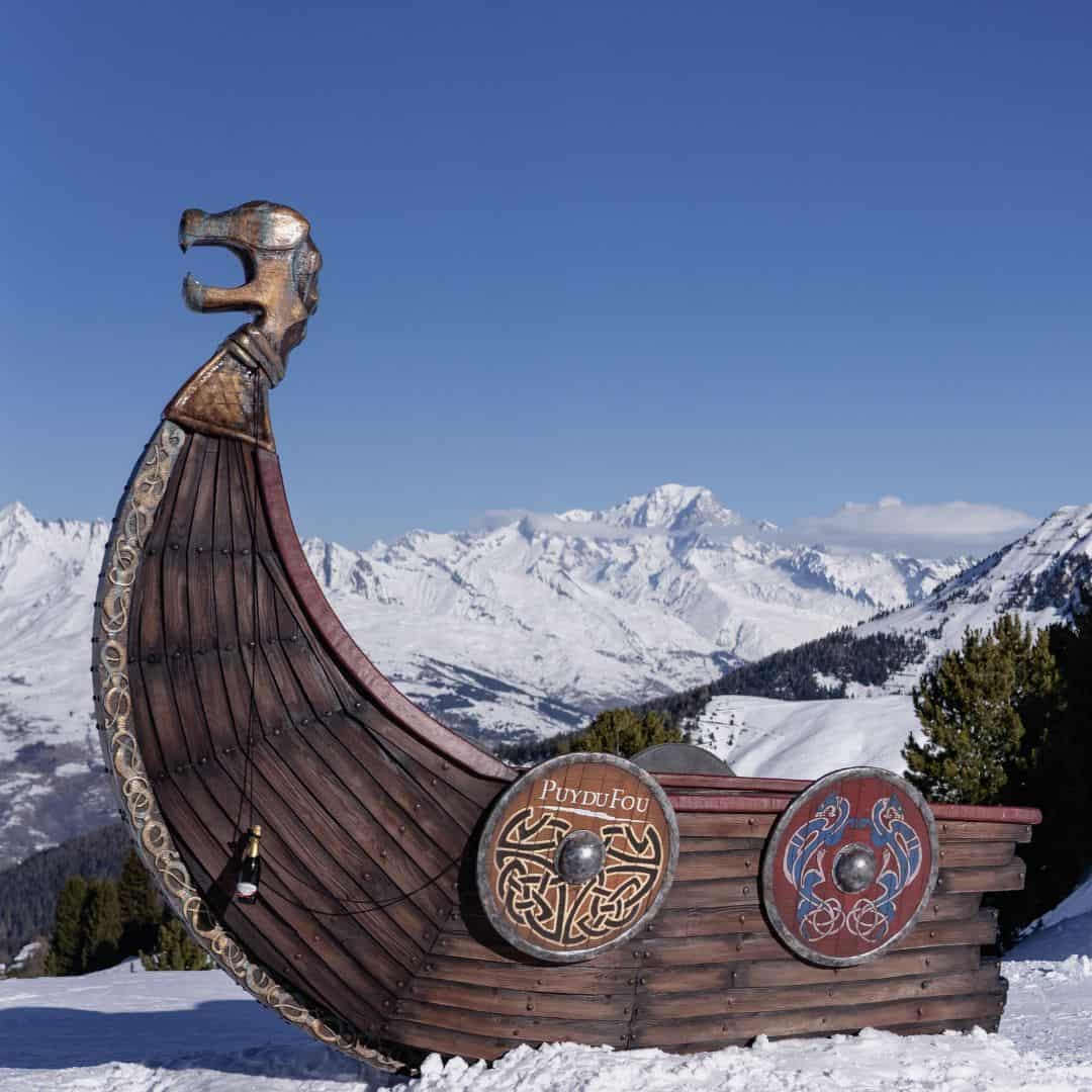 Wooden Viking ship sculpture with intricate carvings and round shields, including one labeled “Puy du Fou,” stands on a snowy mountain slope with stunning views of the French Alps in the background. The clear blue sky and snow-covered peaks create a dramatic alpine setting.