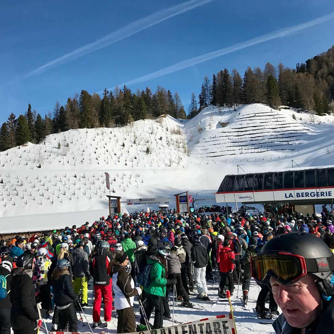Large crowd of skiers and snowboarders queue at the base of the La Bergerie ski lift on a sunny winter day, surrounded by snow-covered slopes and pine trees. The lift area is bustling with colorful ski gear and set against a clear blue sky with visible contrails above.