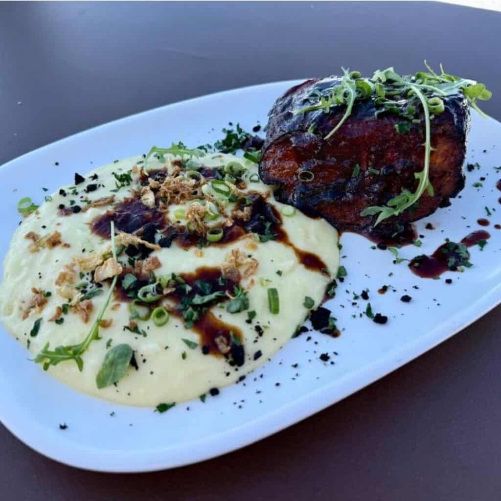 Gourmet plate featuring a glazed, herb-topped cut of meat paired with creamy mashed potatoes, garnished with chopped chives, crispy onions, and a drizzle of dark sauce. Served on a white rectangular plate against a dark tabletop background.