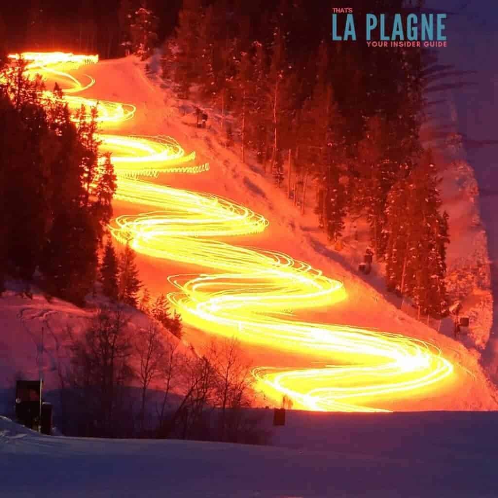 A nighttime ski slope in La Plagne glows with a winding trail of golden lights, captured in a long-exposure photo of a torchlit descent through the snow-covered forest. The illuminated S-curves create a dramatic visual against the dark trees and snowy mountain.