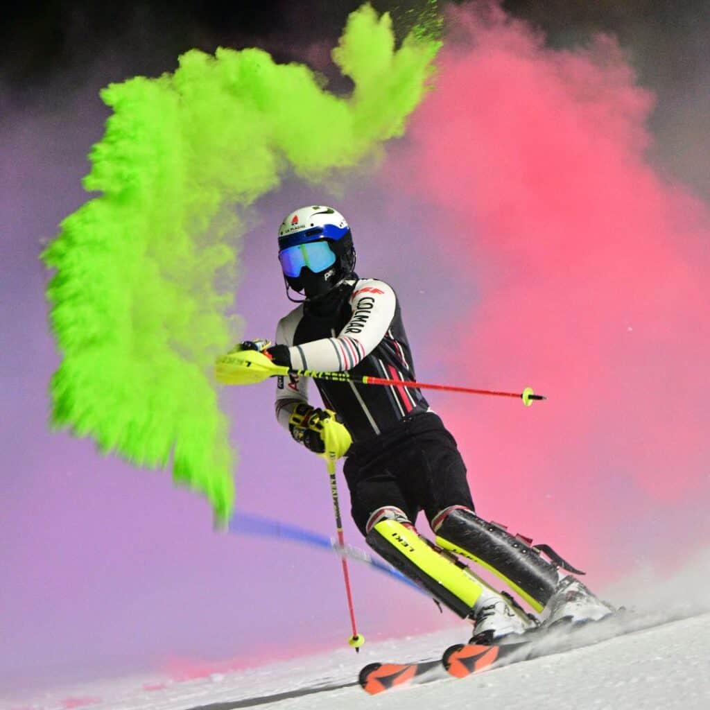 A skier in a black and white racing suit carves through the snow at night, releasing a vibrant burst of green powder from their glove. The background is illuminated with swirling pink and purple smoke, creating a dramatic and colorful effect. The skier's reflective visor and neon yellow gear add to the dynamic, high-energy atmosphere.