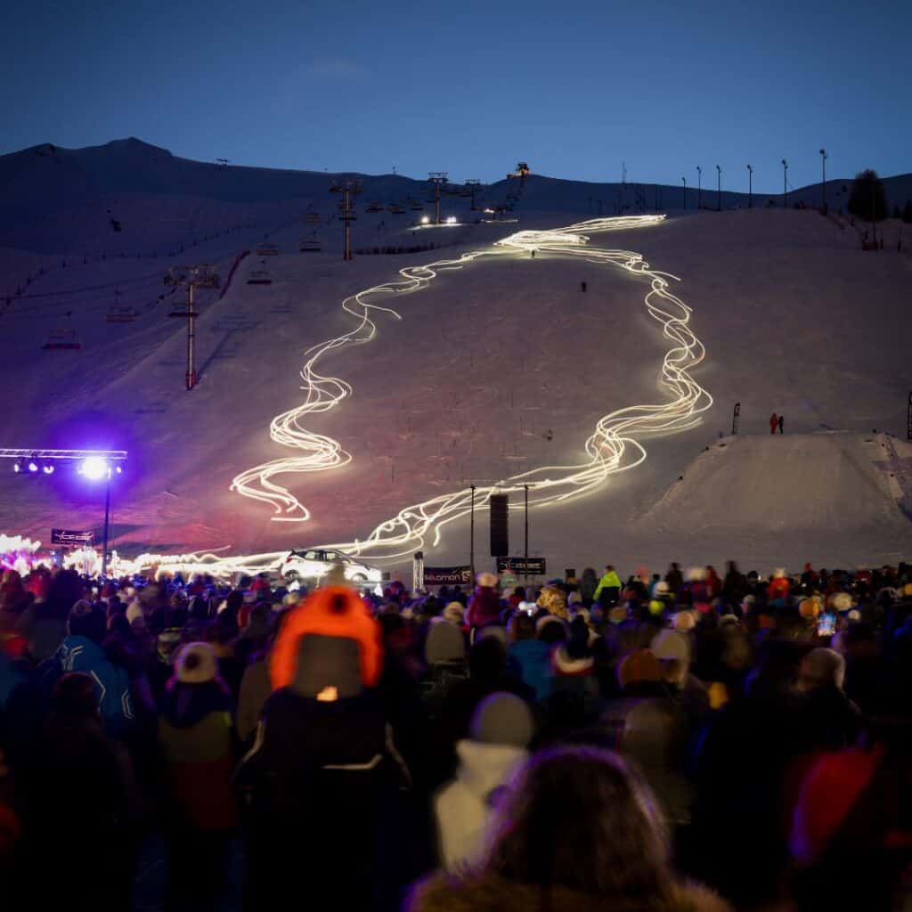 A large crowd gathers at the base of a ski slope at dusk, watching as skiers descend in a mesmerizing torchlight descent formation. The glowing, winding path of lights creates a stunning visual against the snow-covered mountain, with ski lifts and a clear evening sky in the background. The event stage and speakers are illuminated, adding to the lively atmosphere of this nighttime spectacle.
