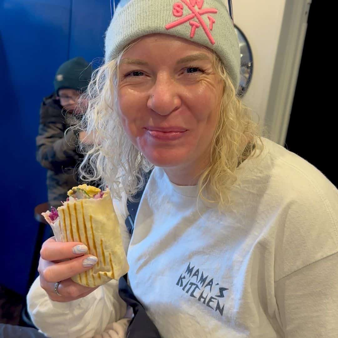 A woman with curly blonde hair, wearing a white "Mama’s Kitchen" sweatshirt and a beanie with pink embroidery, takes a bite of a grilled wrap. She is smiling as she enjoys her meal at Mama’s Kitchen.