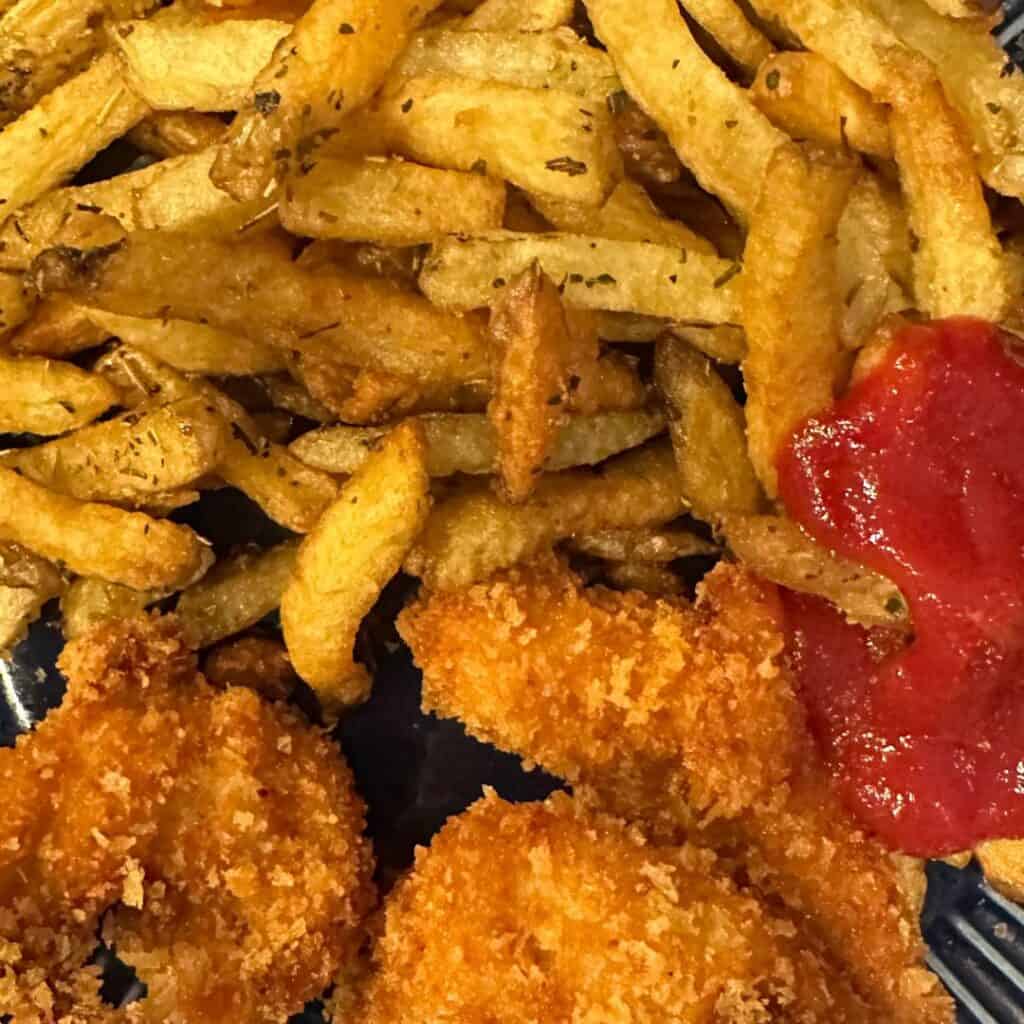 A close-up of a plate at Mama’s Kitchen filled with crispy, golden French fries seasoned with herbs, alongside breaded chicken nuggets. A dollop of ketchup sits on the side of the plate.