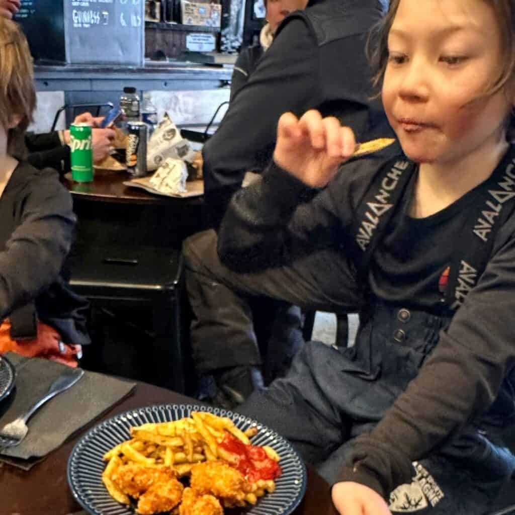 A young child in ski overalls eats a French fry while sitting at a table inside Mama’s Kitchen. A plate of crispy chicken nuggets, fries, and ketchup sits in front of them, while other diners are seen in the background.