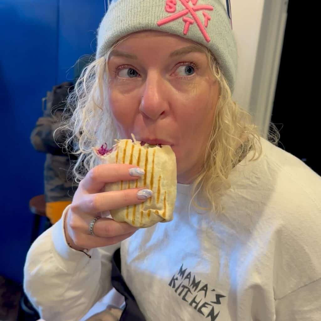 A woman with curly blonde hair, wearing a white "Mama’s Kitchen" sweatshirt and a beanie with pink embroidery, takes a bite of a grilled wrap. She looks off to the side as she enjoys her meal at Mama’s Kitchen.