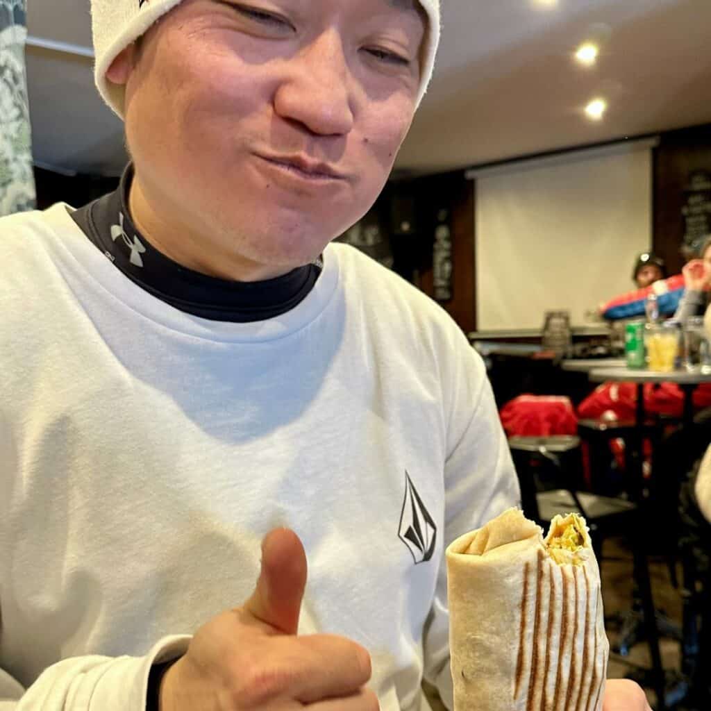 A man in a white long-sleeved shirt and beanie smiles while taking a bite of a grilled wrap at Mama’s Kitchen. He gives a thumbs-up as he enjoys his meal in a cozy restaurant setting with other diners in the background.