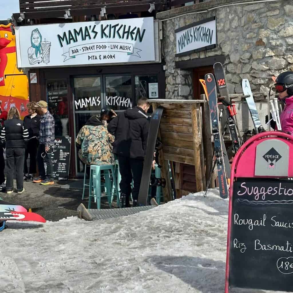 A lively outdoor scene at Mama’s Kitchen, a rustic-style restaurant with a wooden and stone exterior. People in ski gear gather outside, some chatting and others enjoying food, while snowboards and skis are propped up nearby. A chalkboard menu displays the day's special: "Rougail Saucisse" with "Riz Basmati" for 18€.