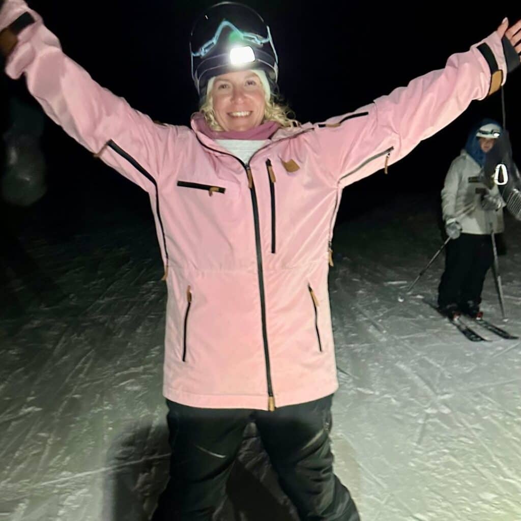 A woman in a pink ski jacket and helmet with a headlamp smiles with her arms wide open during a nighttime ski adventure. The dimly lit snowy slope behind her features another skier preparing for the descent.