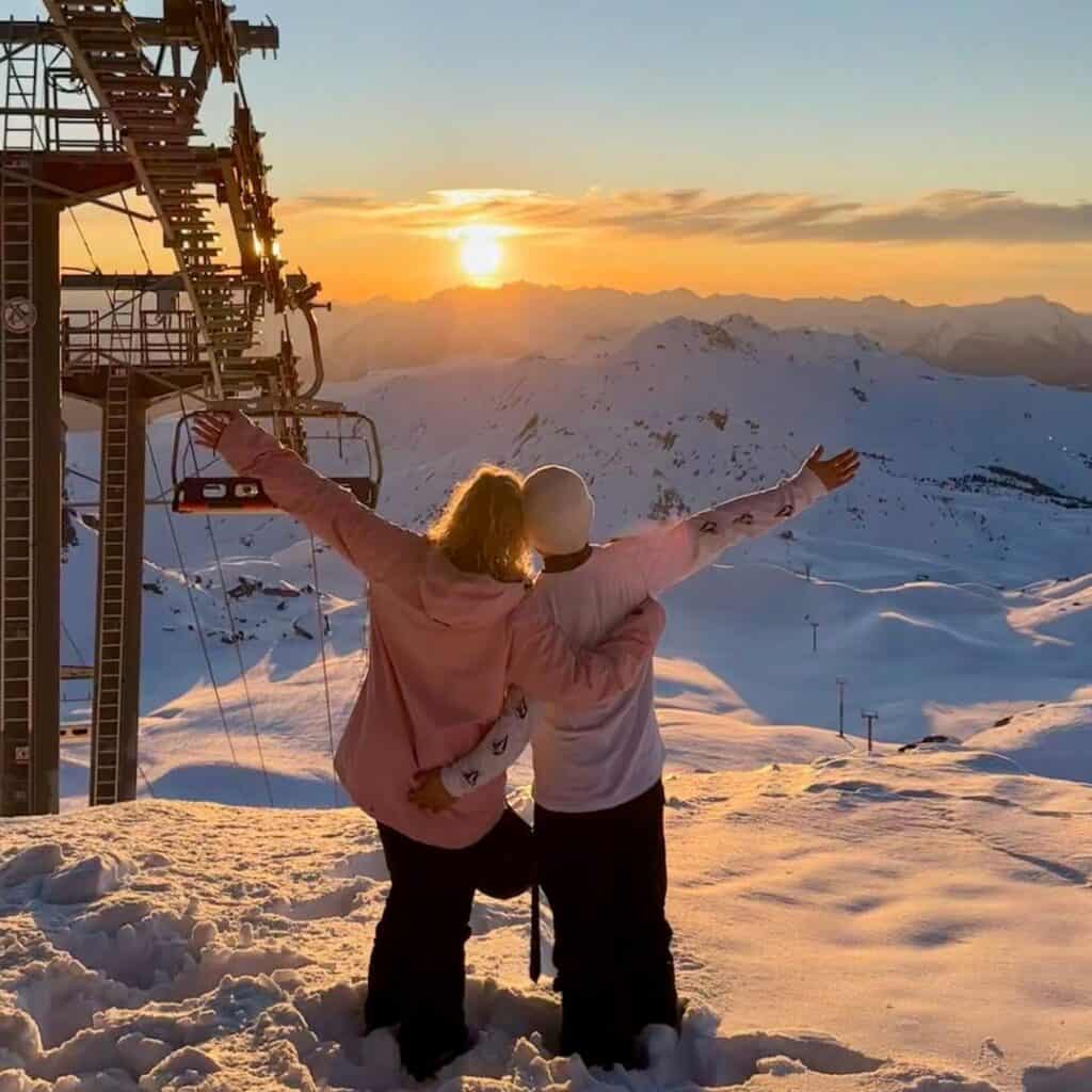 A couple stands in the snow on a mountain, arms outstretched, embracing a breathtaking sunset over snow-covered peaks. They are dressed in winter gear, with the person on the left wearing a pink jacket and the one on the right in a white beanie and matching top. A ski lift structure is visible to their left, adding to the alpine atmosphere of this serene, golden-lit scene.