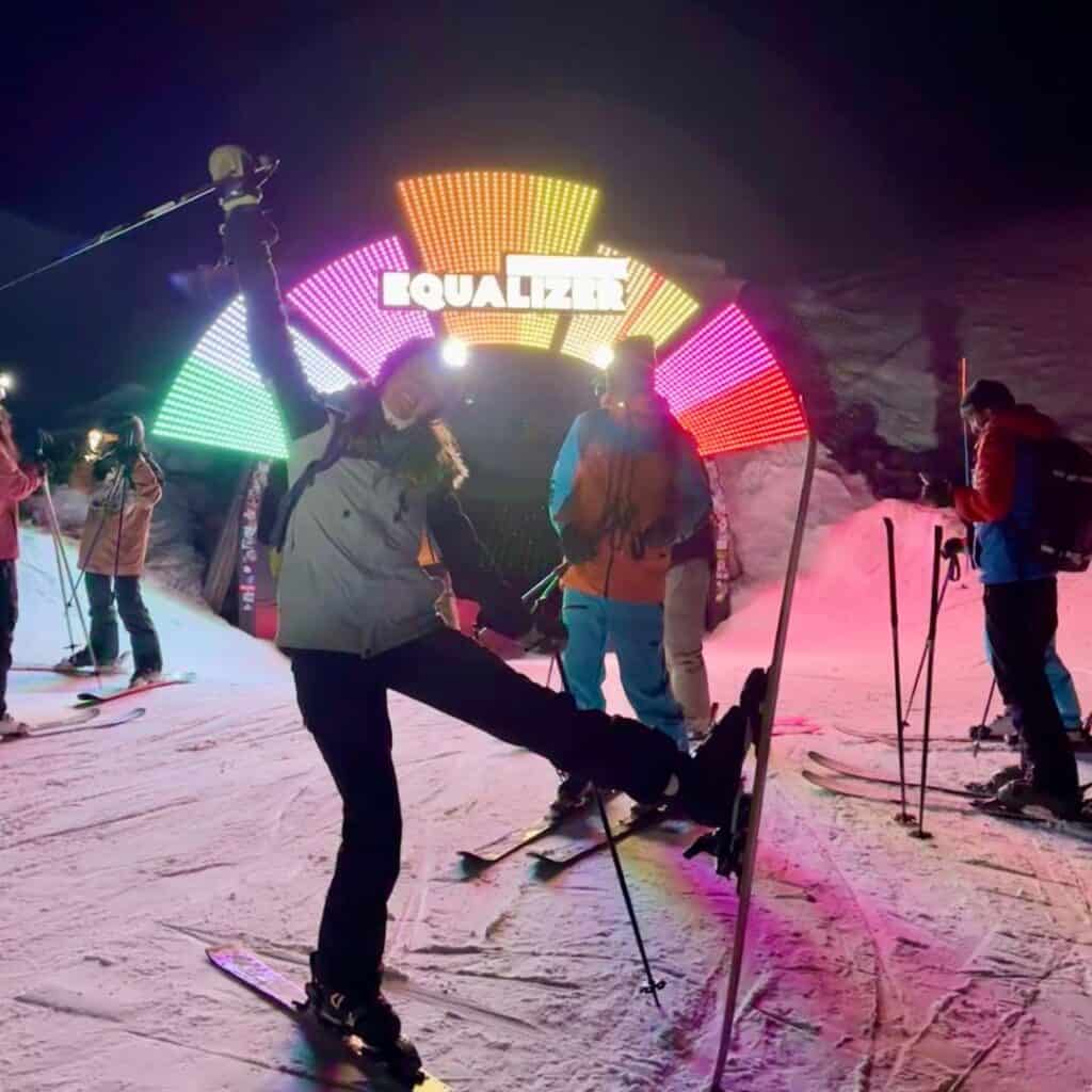 A skier poses playfully in front of a colorful, illuminated 'EQUALIZER' sign at a nighttime ski event. Other skiers, dressed in warm winter gear, gather around the glowing archway, enjoying the lively atmosphere.