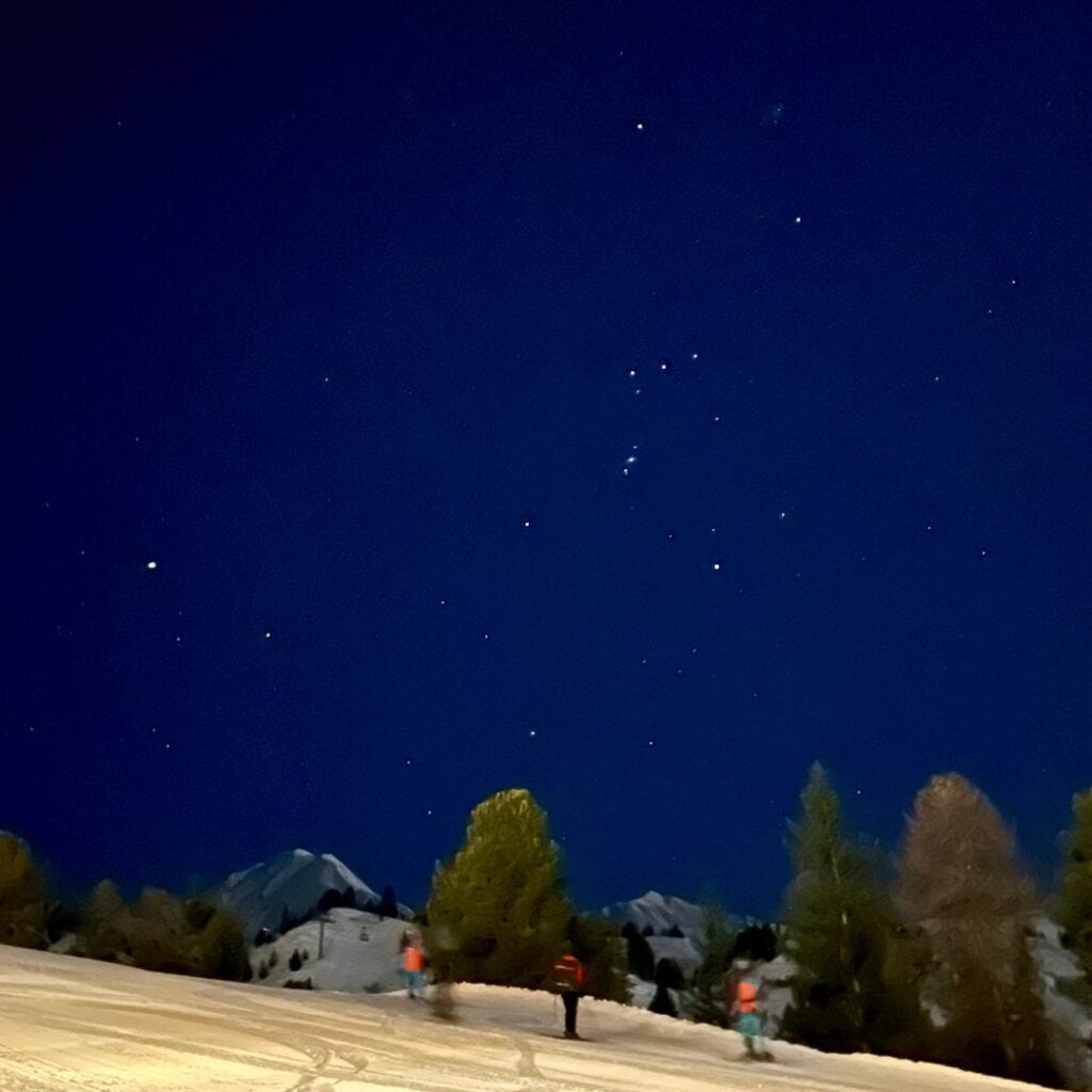 A stunning night sky filled with stars shines above a snowy mountain landscape. Silhouettes of skiers and snow-covered trees stand beneath the celestial view, creating a peaceful winter scene.