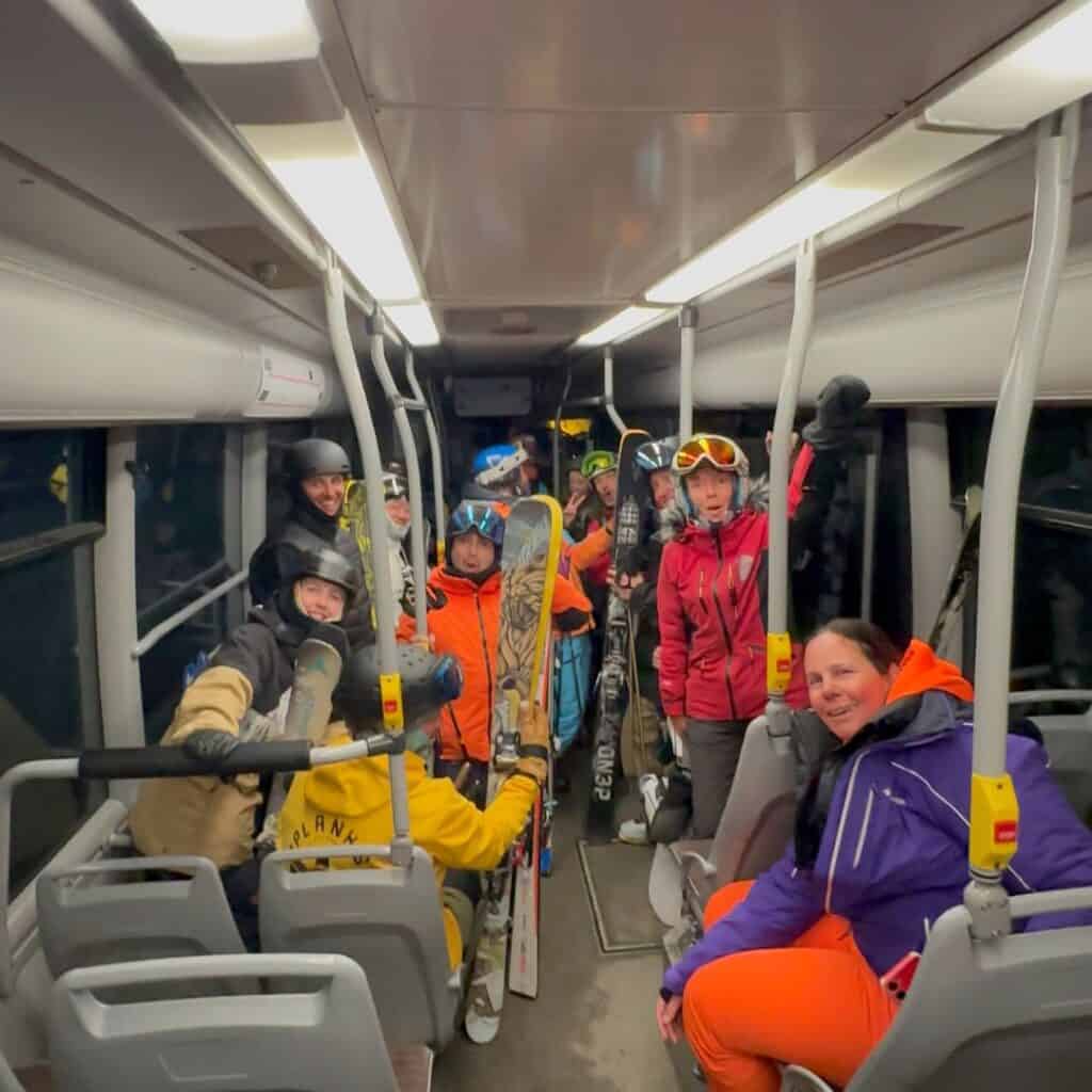 A group of skiers and snowboarders, still in their winter gear, pack into a public bus after a long day on the slopes. Their faces show excitement and exhaustion as they hold onto ski poles and snowboards while chatting with each other.