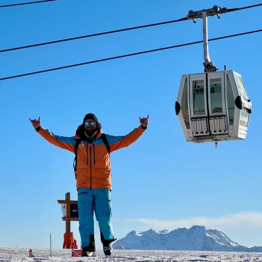 A man wearing an orange and blue ski jacket, sunglasses, and a beanie stands on a snowy mountaintop, raising his arms in a celebratory pose. A ski gondola hovers above him on the cable, with a backdrop of clear blue skies and snow-capped peaks