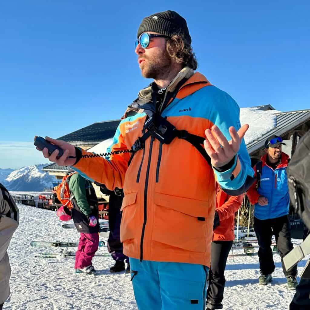 A ski guide wearing an orange and blue jacket, sunglasses, and a black beanie gestures while speaking into a handheld radio. Other skiers in colorful gear stand behind him near a mountain hut, with skis scattered in the snow and a bright blue sky overhead.