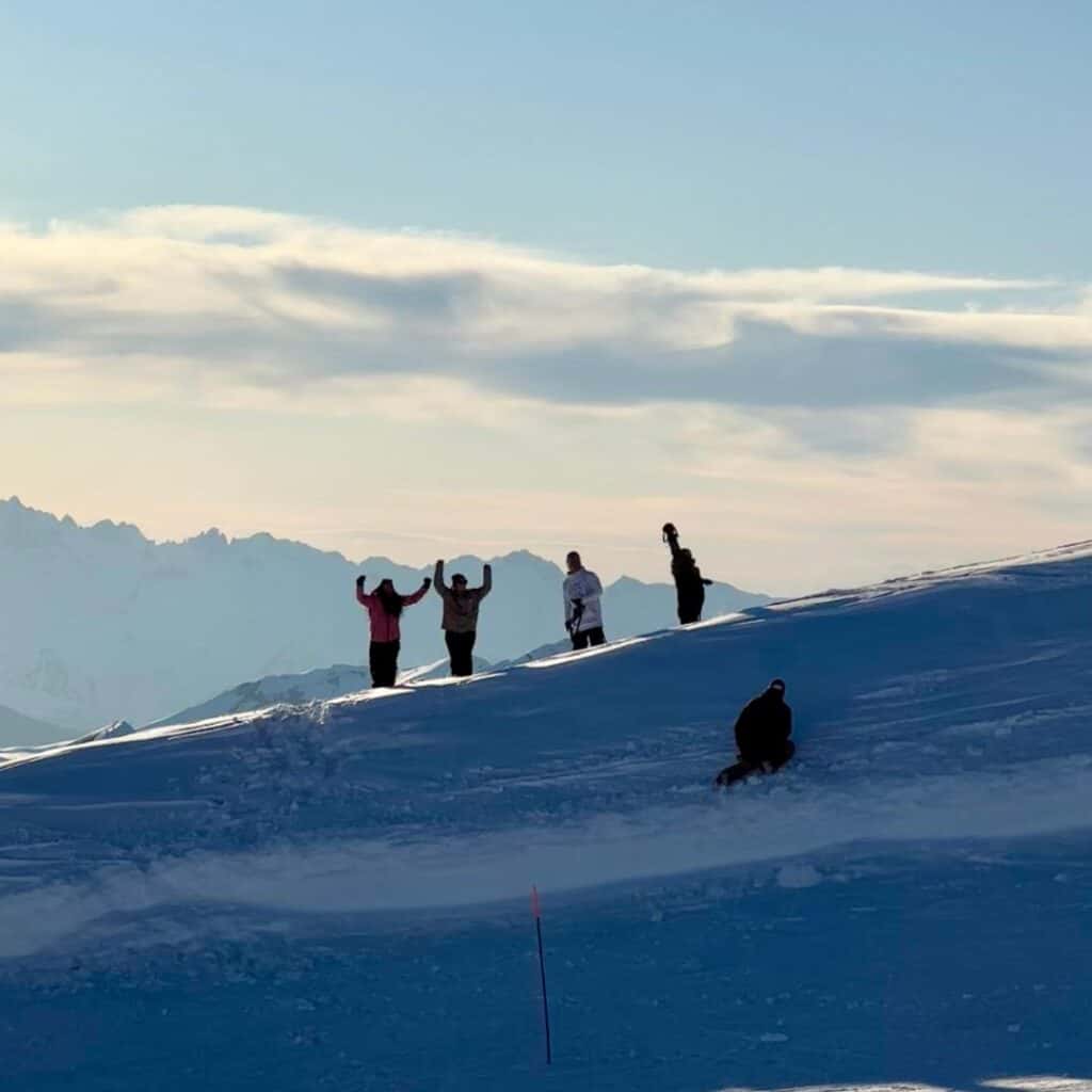 Four people stand atop a snow-covered ridge, silhouetted against a soft, cloudy sky, raising their arms in excitement. Below them, another person kneels in the snow, possibly adjusting their gear or preparing to snowboard