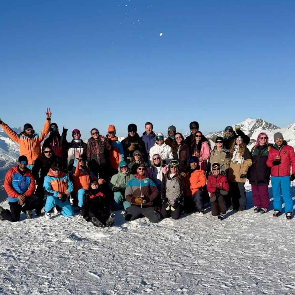 A large group of people in colorful ski jackets, goggles, and winter gear pose together on a snowy mountaintop, smiling under a clear blue sky. Some throw snow into the air, while snow-covered peaks stretch behind them in the distance.