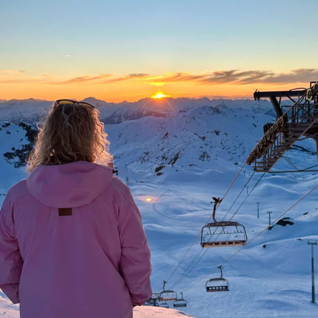 A woman in a pink jacket stands with her back to the camera, gazing at a breathtaking mountain sunset. Snow-covered slopes stretch below, with ski lifts suspended in the golden light of the setting sun.
