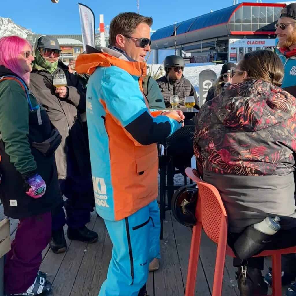 A ski instructor in an orange and blue jacket interacts with a group of people at an outdoor après-ski gathering. The scene is lively, with skiers in colorful gear enjoying drinks on a wooden deck, surrounded by snowy peaks.