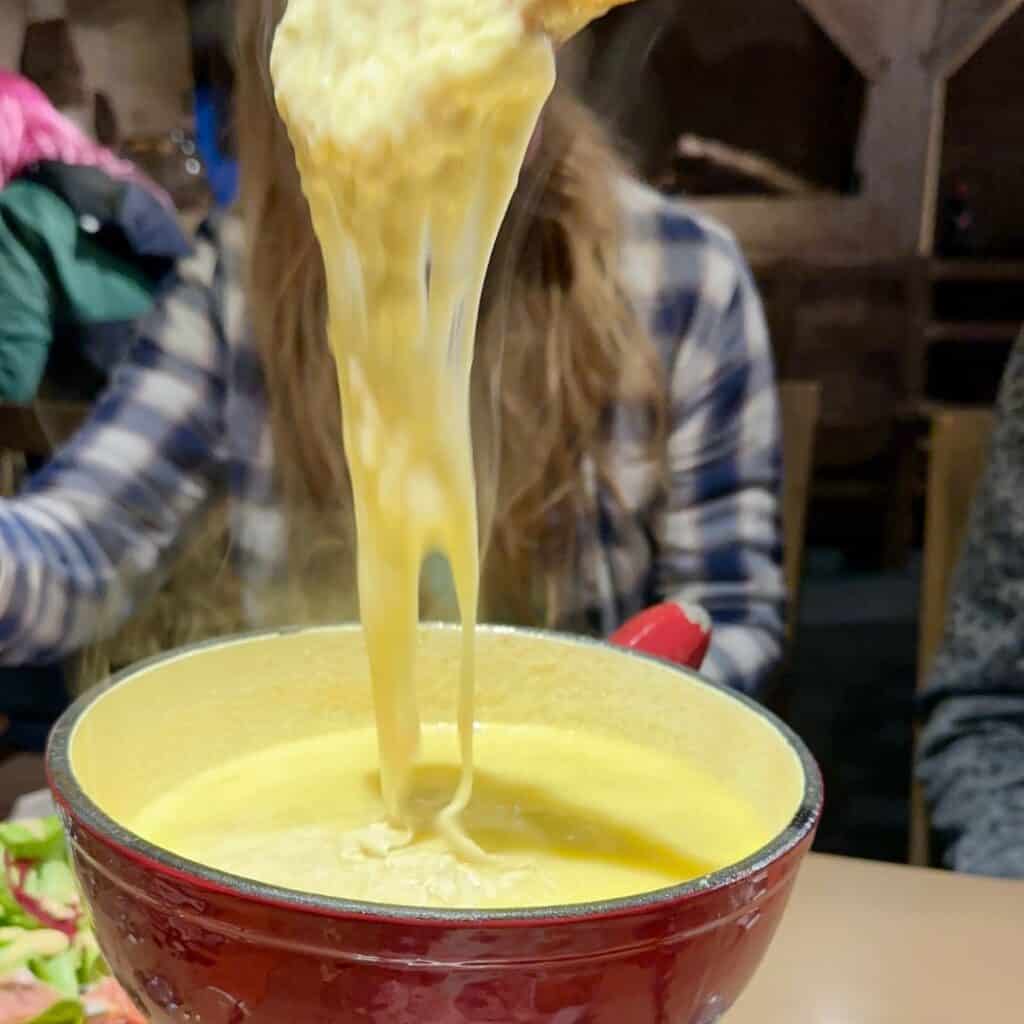 A steaming pot of cheese fondue sits on the table as a person lifts a long, gooey strand of melted cheese with a piece of bread. The warm, golden cheese contrasts with the cozy, wooden interior of the restaurant.