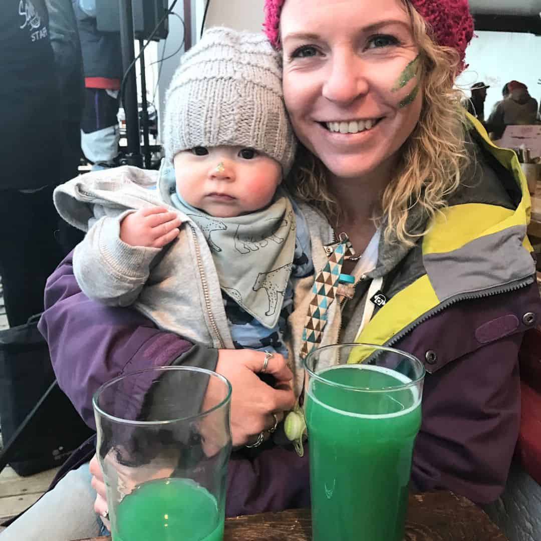 A smiling woman wearing a pink knit beanie and a colorful winter jacket holds a baby dressed in a gray beanie and a patterned bandana bib. The baby looks curiously at the camera with a small smudge of green on their nose. In front of them on a wooden table are two tall glasses of vibrant green beer, likely for St. Patrick’s Day celebrations. The background shows an indoor gathering with other people dressed in warm clothing.