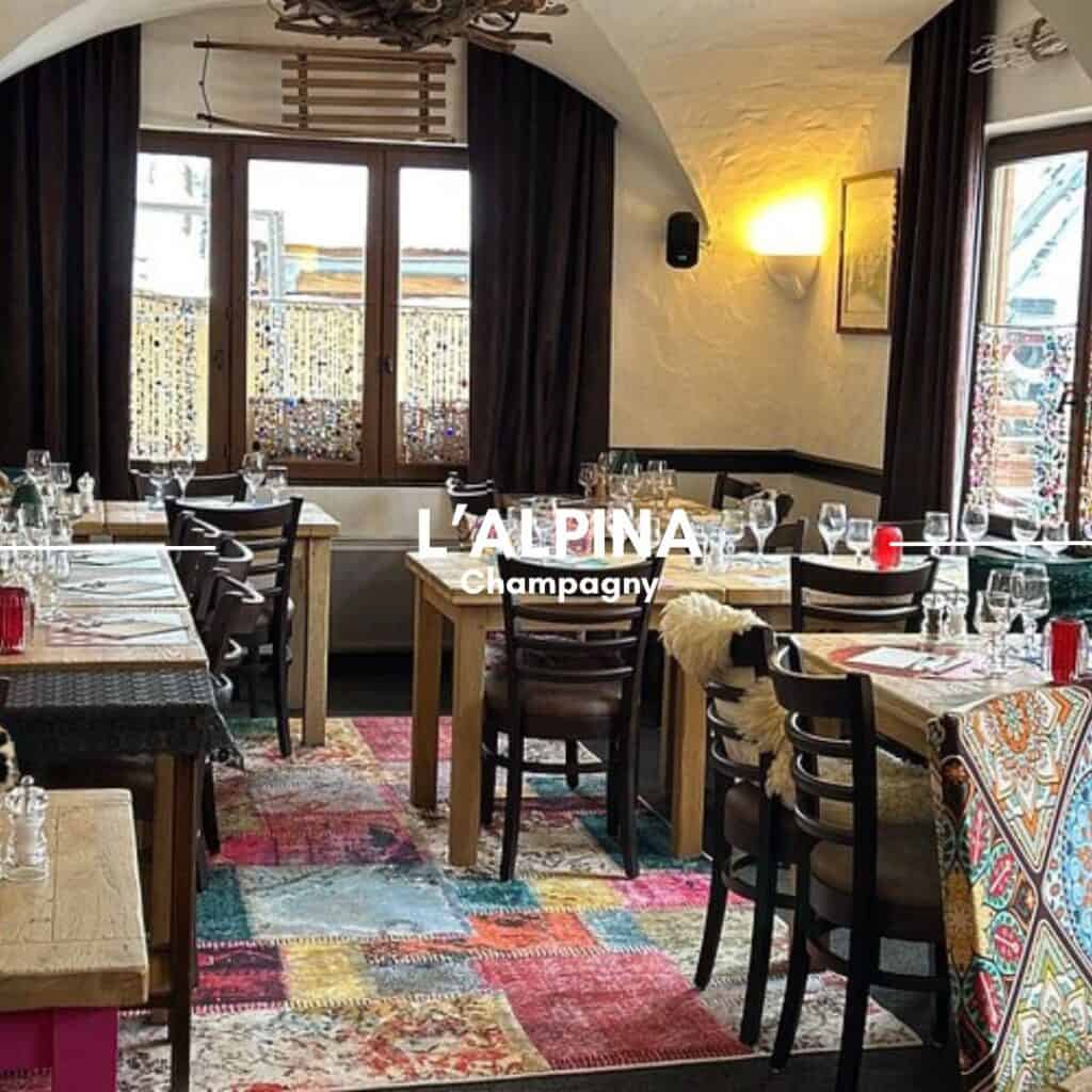 L'ALPINA, Champagny – A cozy alpine restaurant with a rustic yet elegant interior, featuring wooden tables set with wine glasses, decorative rugs, and warm lighting. The large windows provide a glimpse of the snowy outdoors.