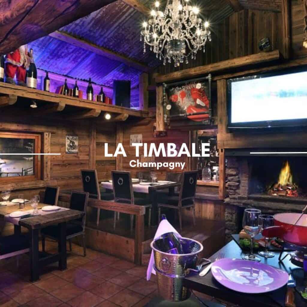 La Timbale restaurant in Champagny features a rustic yet elegant wooden interior with a roaring fireplace, a crystal chandelier, and dim purple lighting. The space is decorated with vintage ski-themed artwork, a mounted TV, and a mezzanine stocked with wine bottles. A fondue set and a champagne bucket sit on a table, adding to the cozy alpine dining experience.