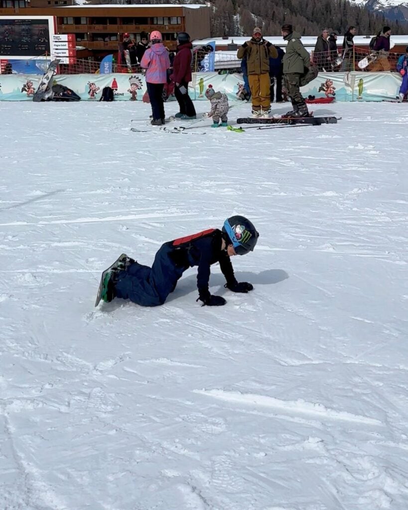 A child in dark snow gear crawls on the snow with a snowboard attached, capturing a learning moment on the slopes at La Plagne. Onlookers and skiers watch in the distance.