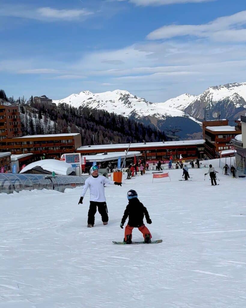 A snowboarding instructor gestures to a child on a snowboard at La Plagne, with scenic snow-covered mountains and chalet-style buildings in the background. The scene captures a lesson in progress with other skiers nearby.