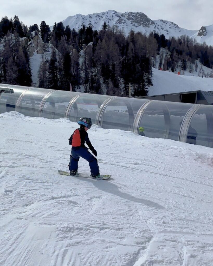 A young snowboarder in a navy snowsuit rides down a gentle slope at La Plagne, next to a transparent tunnel lift and surrounded by pine trees and rugged alpine peaks. The child maintains balance while carving through the snow.