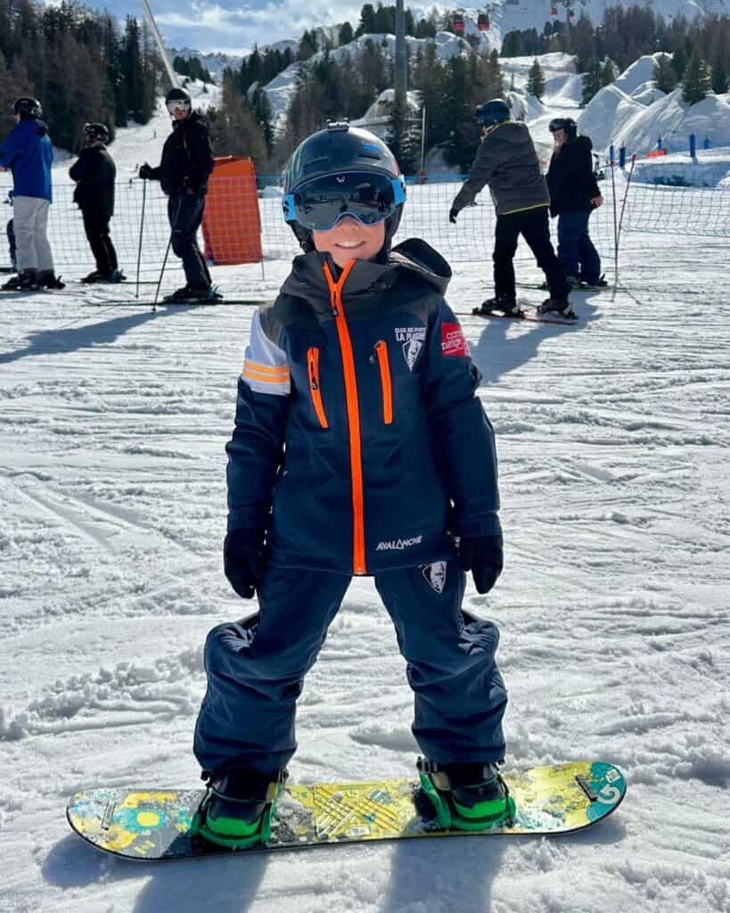 A smiling child in full snowboarding gear poses confidently on their board under sunny skies at La Plagne. Other skiers line up in the background near a netted slope area, with ski lifts and snowy trees in the distance.
