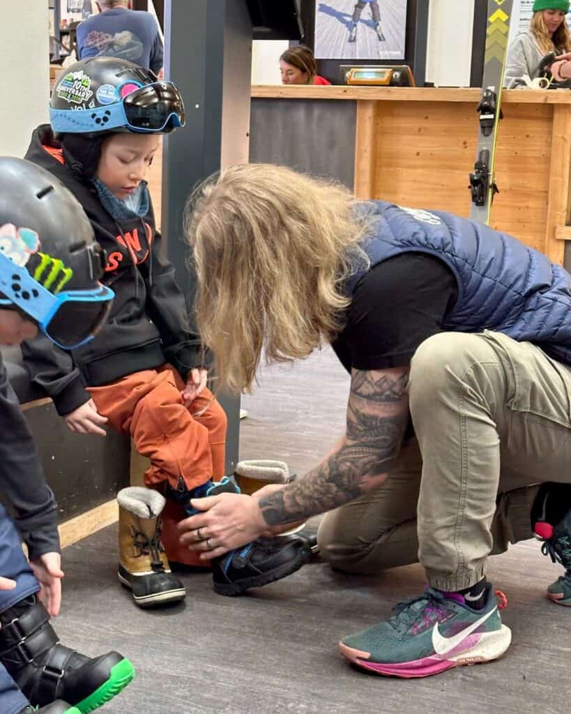 A snowboard instructor helps a young child fasten snowboarding boots indoors at La Plagne, as other kids wait patiently nearby. The setting is cozy and filled with ski rental gear.