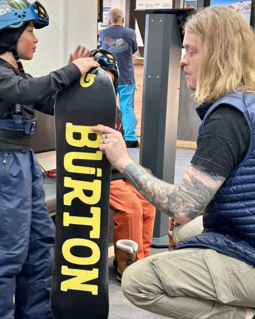 A young boy in a helmet and snow gear receives a snowboarding lesson indoors as the instructor points to a Burton snowboard. This image captures a teaching moment at La Plagne’s snowboarding school.