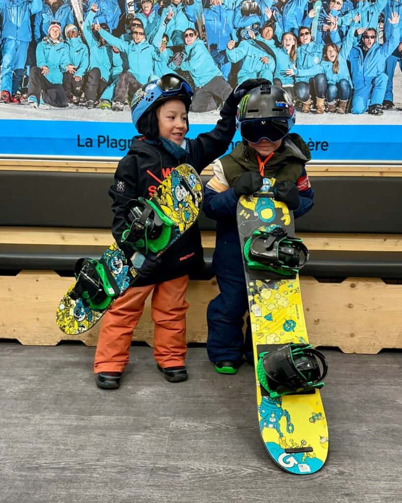 Two kids in snowboarding gear pose with colorful snowboards in front of a large group photo of La Plagne instructors. One child playfully ruffles the other’s helmet, adding a fun vibe to the scene.