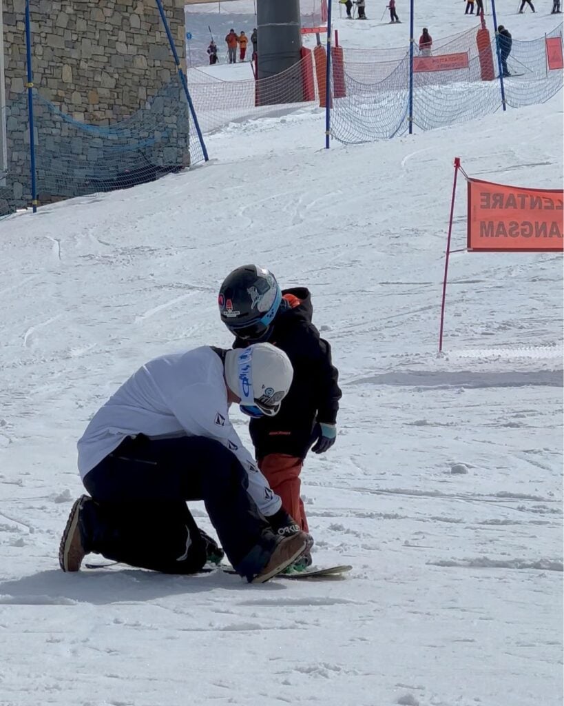 An instructor kneels on the snow to adjust a child's snowboard bindings at La Plagne, preparing for a lesson. The kid stands still in full snowboarding gear under clear skies.