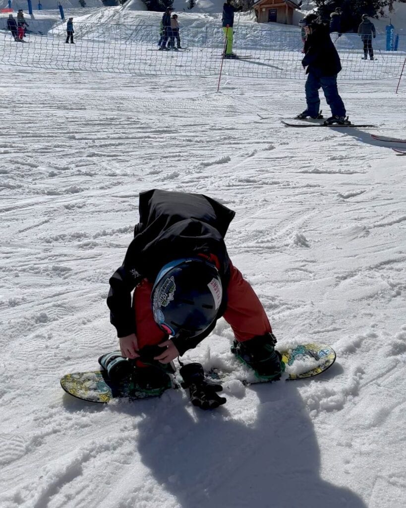 A young child in orange snow pants crouches in the snow at La Plagne, strapping into a bright green snowboard. Other kids snowboard nearby under sunny skies.