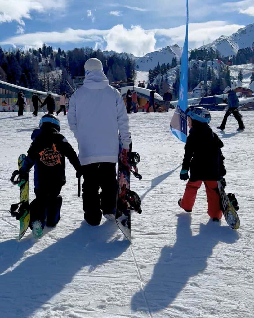 A snowboard instructor walks hand in hand with two snowboarding kids at La Plagne, all carrying their boards toward the mountain. Snow-covered peaks and ski lifts fill the background.