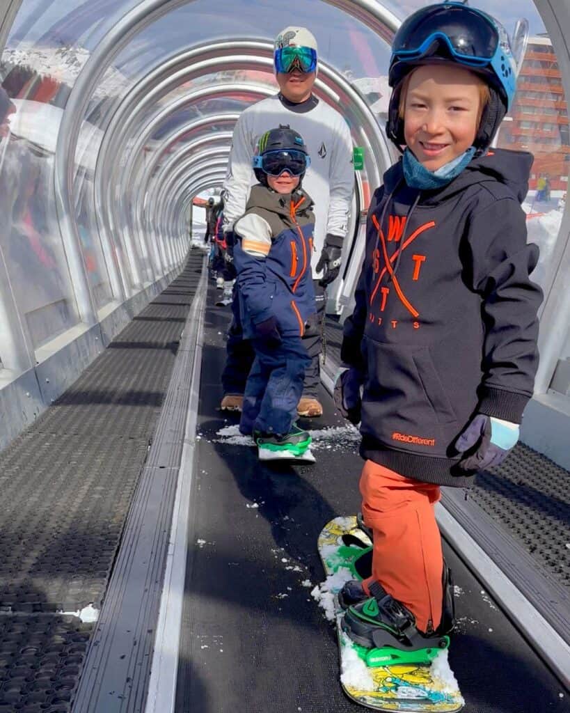 Three snowboarding kids and an adult ride a covered conveyor lift at a snowy La Plagne resort, smiling and ready to hit the slopes. Their snowboards and gear reflect bright colors and excitement.