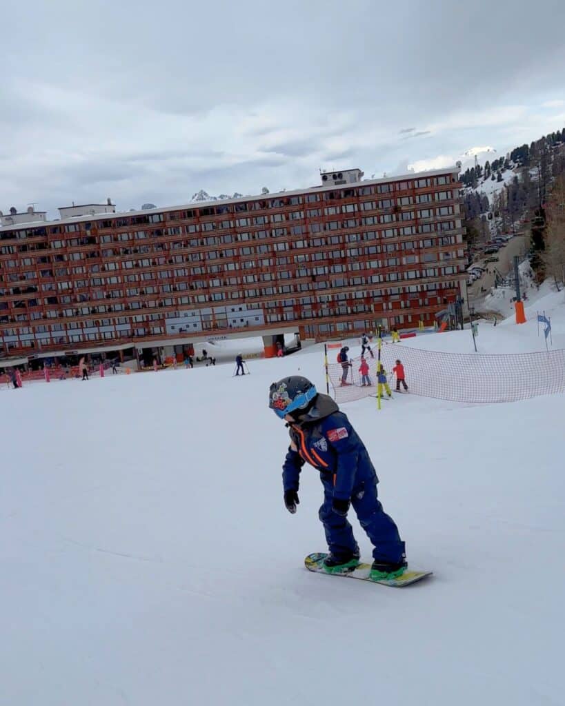 A child glides down a snowy slope at La Plagne on a snowboard, with a large chalet-style hotel in the background. The child appears confident and focused during the ride.