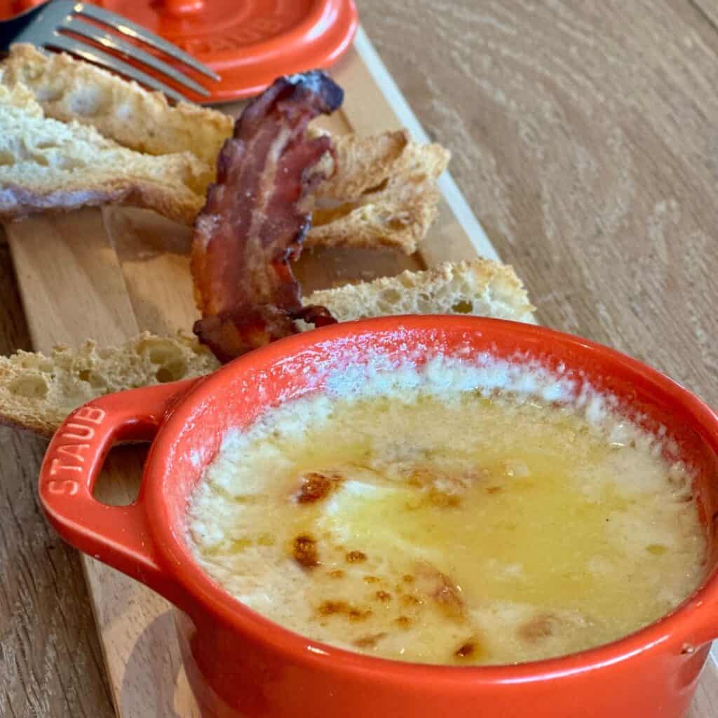 A bubbling dish of melted cheese served in a rustic red Staub cocotte, accompanied by crispy bread slices and a strip of bacon, all placed on a wooden board. At Le Chairlift restaurant, La Plagne.