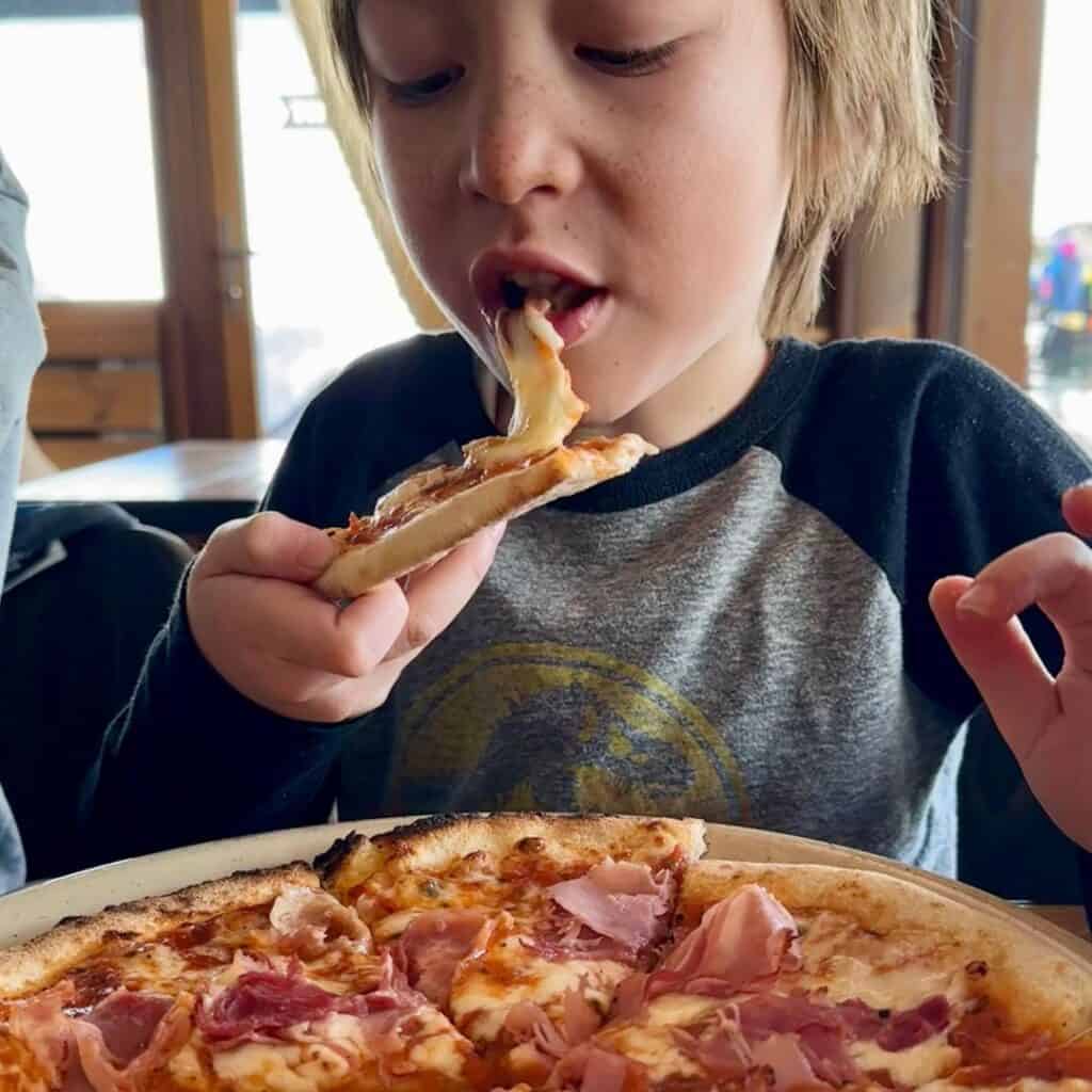 A young child with light skin and freckles enjoying a slice of pizza with ham, stretching a piece of gooey melted cheese as they take a bite. At Le Chairlift restaurant, La Plagne.