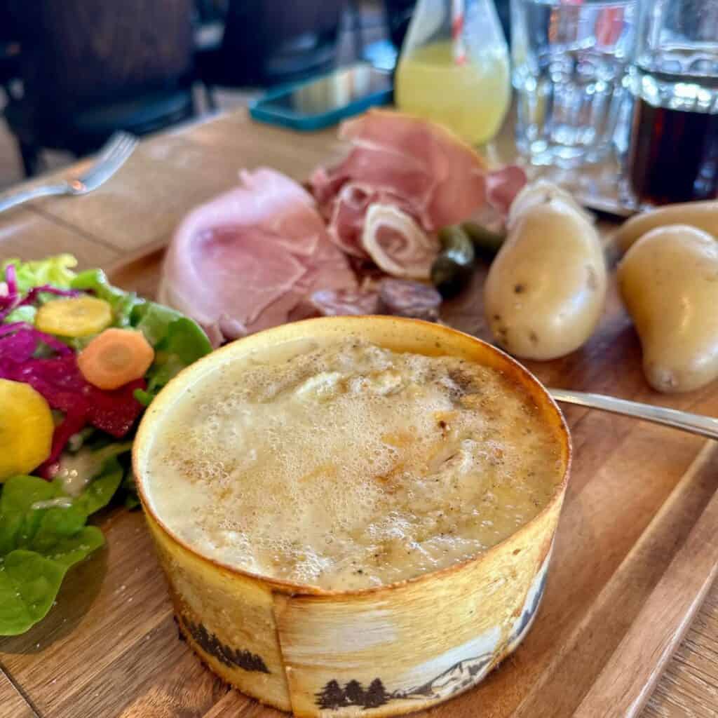 A traditional French raclette meal featuring a wheel of melted cheese in a wooden dish, served with boiled potatoes, charcuterie, and a fresh side salad. At Le Chairlift restaurant, La Plagne.