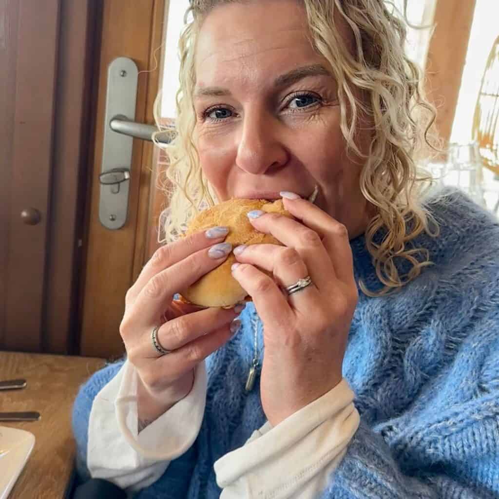 A woman with curly blonde hair and blue eyeshadow smiling as she takes a bite of a sandwich at Le Chairlift restaurant, with a cozy wooden interior in the background. At Le Chairlift restaurant, La Plagne.