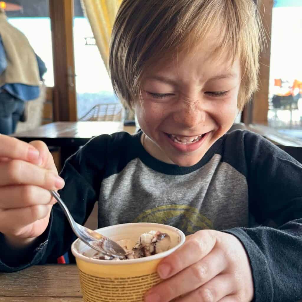 A young child laughing while enjoying a cup of chocolate-swirled ice cream, holding a spoon with excitement. At Le Chairlift restaurant, La Plagne.