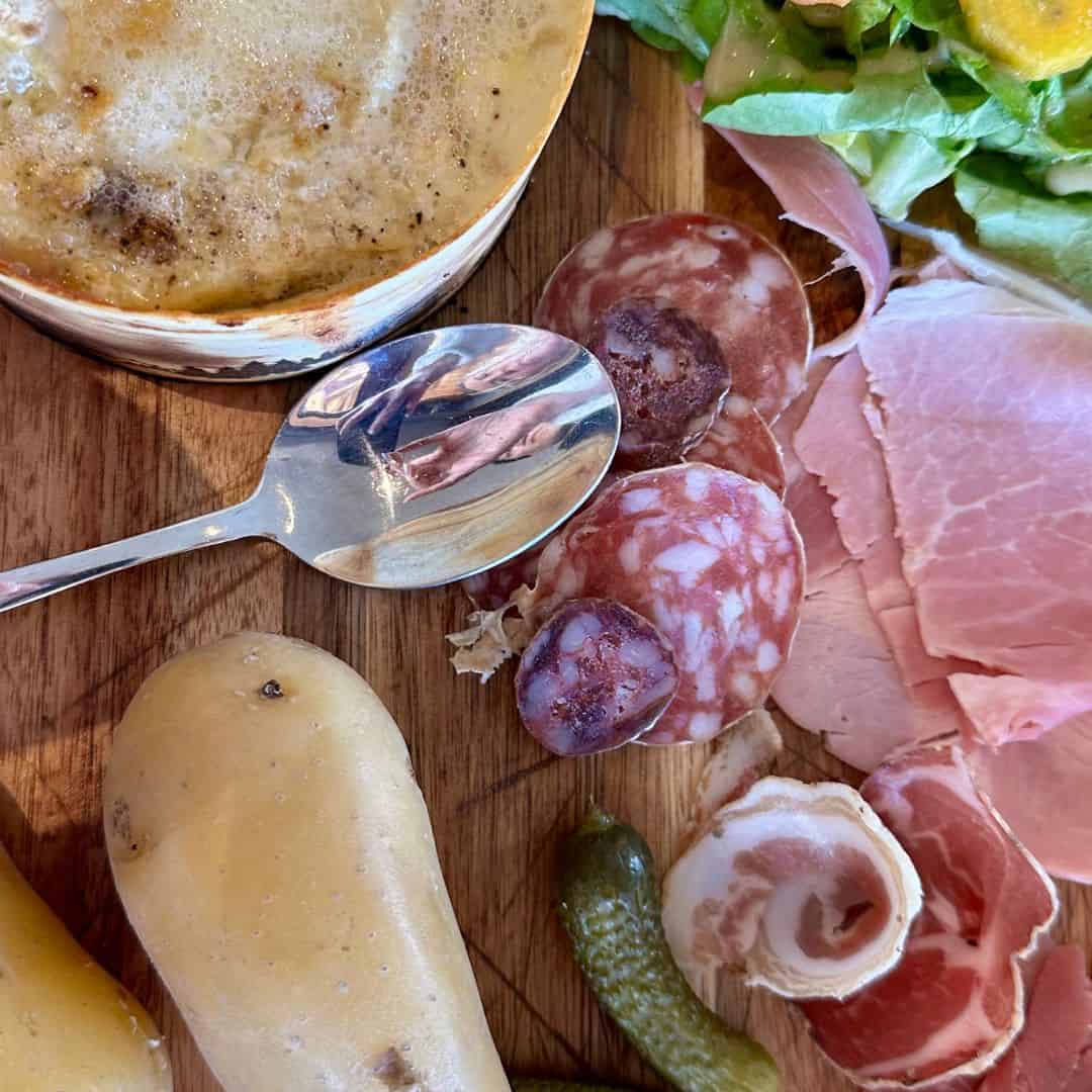 A close-up of a charcuterie board with an assortment of cured meats, a bubbling pot of melted cheese, boiled potatoes, pickles, and a silver spoon reflecting the photographer. At Le Chairlift restaurant, La Plagne.
