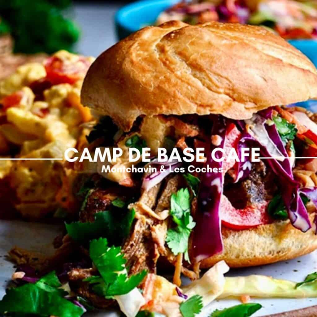 A mouthwatering pulled pork sandwich topped with fresh herbs, red cabbage, and crispy vegetables, served with a side of cheesy potatoes. The vibrant colors and textures highlight the hearty alpine meal. The text overlay reads "Camp de Base Café, Montchavin & Les Coches."